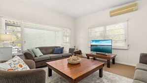A living room with a sofa, coffee table, TV, and large windows with blinds. - Huskisson Delight! by Experience Jervis Bay (Huskisson)