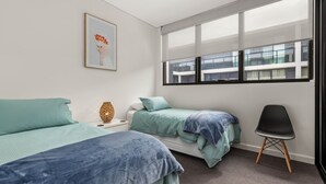 Ein Schlafzimmer mit zwei Betten, einem Sessel, einem Nachttisch mit Lampe und einem Fenster mit Blick auf Gebäude.