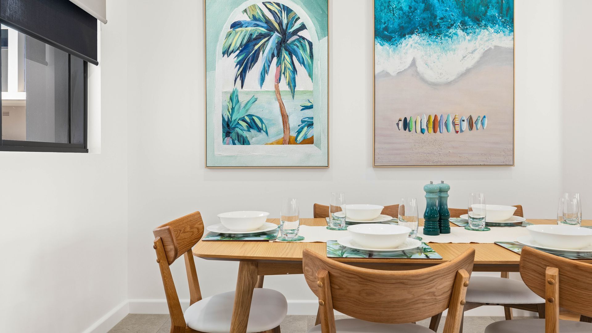 Un comedor con una mesa de madera dispuesta para cuatro personas, rodeada de sillas de madera. Dos cuadros enmarcados cuelgan de la pared sobre la mesa.
