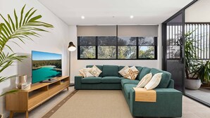 A modern living room with a green sofa, a flat-screen TV, and a large window with a view of trees. - HuskySun by Experience Jervis Bay (Huskisson)