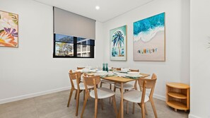 A dining area with a wooden table set for six, surrounded by six chairs. There are three paintings on the wall, a window with a view of buildings, and a small wooden shelf. - HuskySun by Experience Jervis Bay (Huskisson)