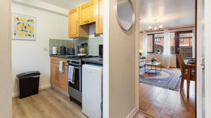 Cocina pequeña con armarios de madera, estufa y refrigerador. Hay un cubo de basura y una mesita con una silla.