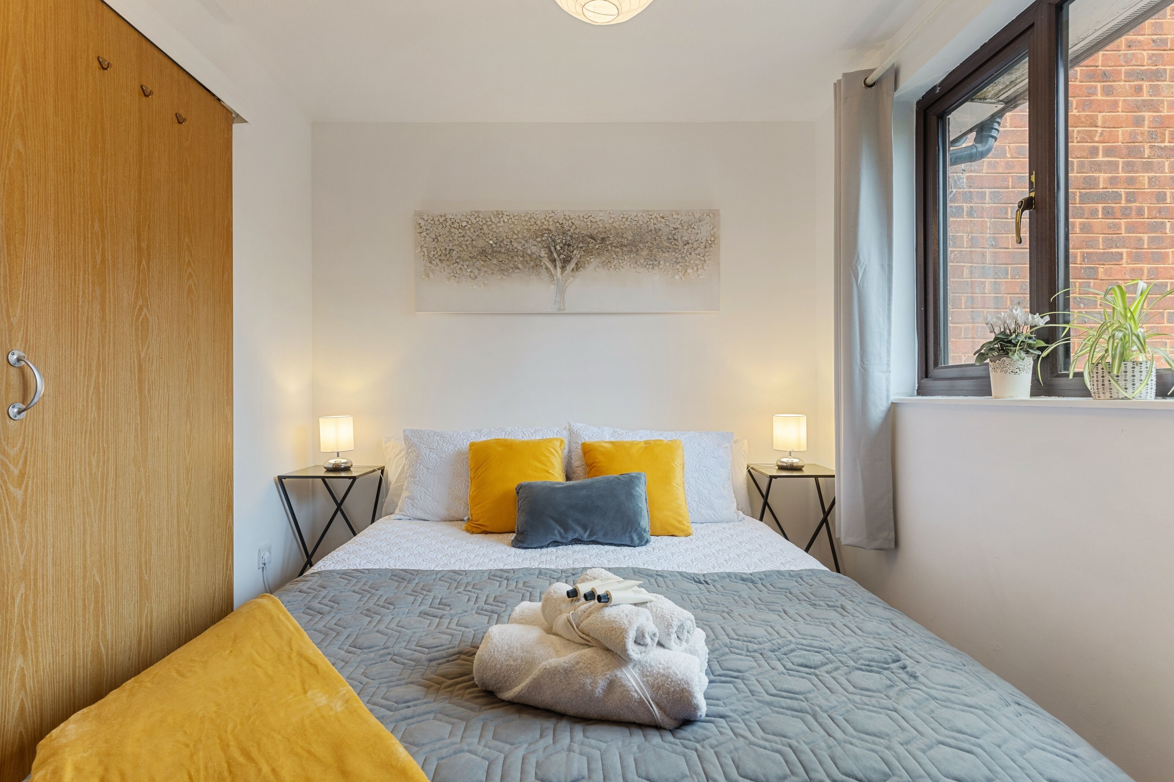A neatly arranged hotel room with a bed, two bedside tables, a window with plants, and a wooden wardrobe.