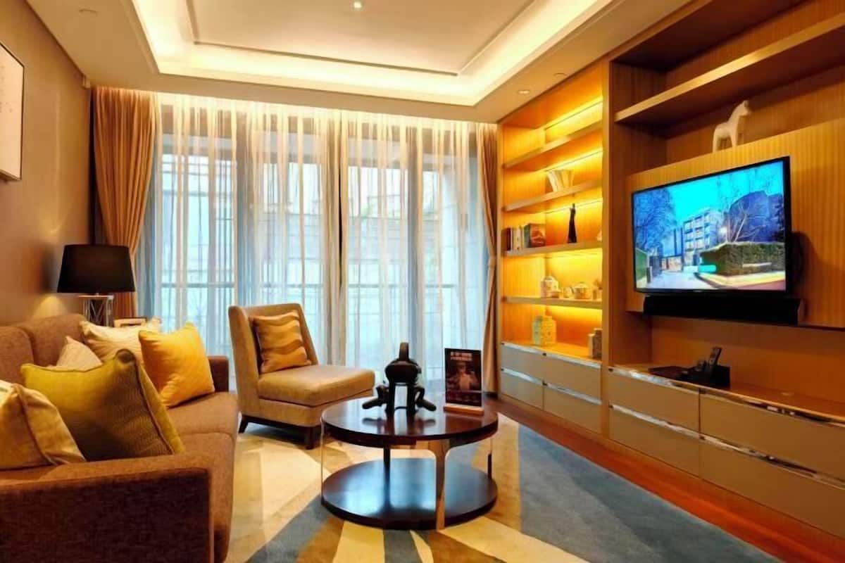 A modern living room with a sofa, armchair, and coffee table, a TV mounted on the wall, and a bookshelf with decorative items.