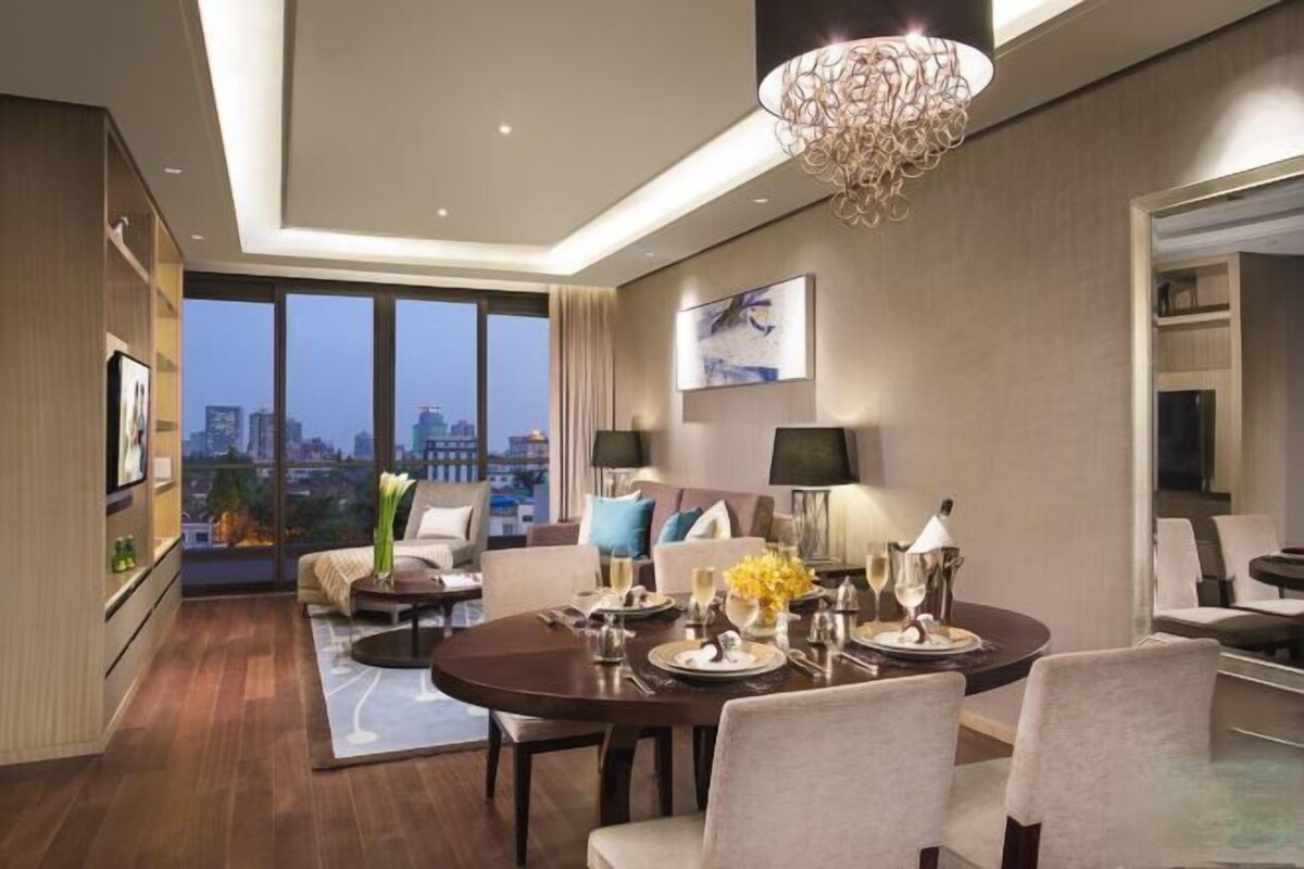 A modern hotel room with a dining area, large windows, a chandelier, and a city view.