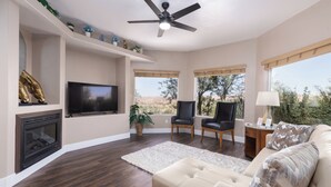 Ein Wohnzimmer mit Kamin, Fernseher, Deckenventilator und großen Fenstern mit Blick ins Freie.