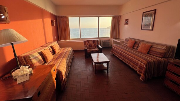 A hotel room with a large window, two sofas, a wooden table, and a lamp. - Islas del Sol (Mazatlán)