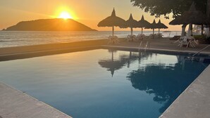 A swimming pool with a view of the sunset over the ocean, surrounded by lounge chairs and thatched umbrellas. - Islas del Sol (Mazatlán)