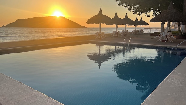 A swimming pool with a view of the sunset over the ocean, surrounded by lounge chairs and thatched umbrellas. - Islas del Sol (Mazatlán)