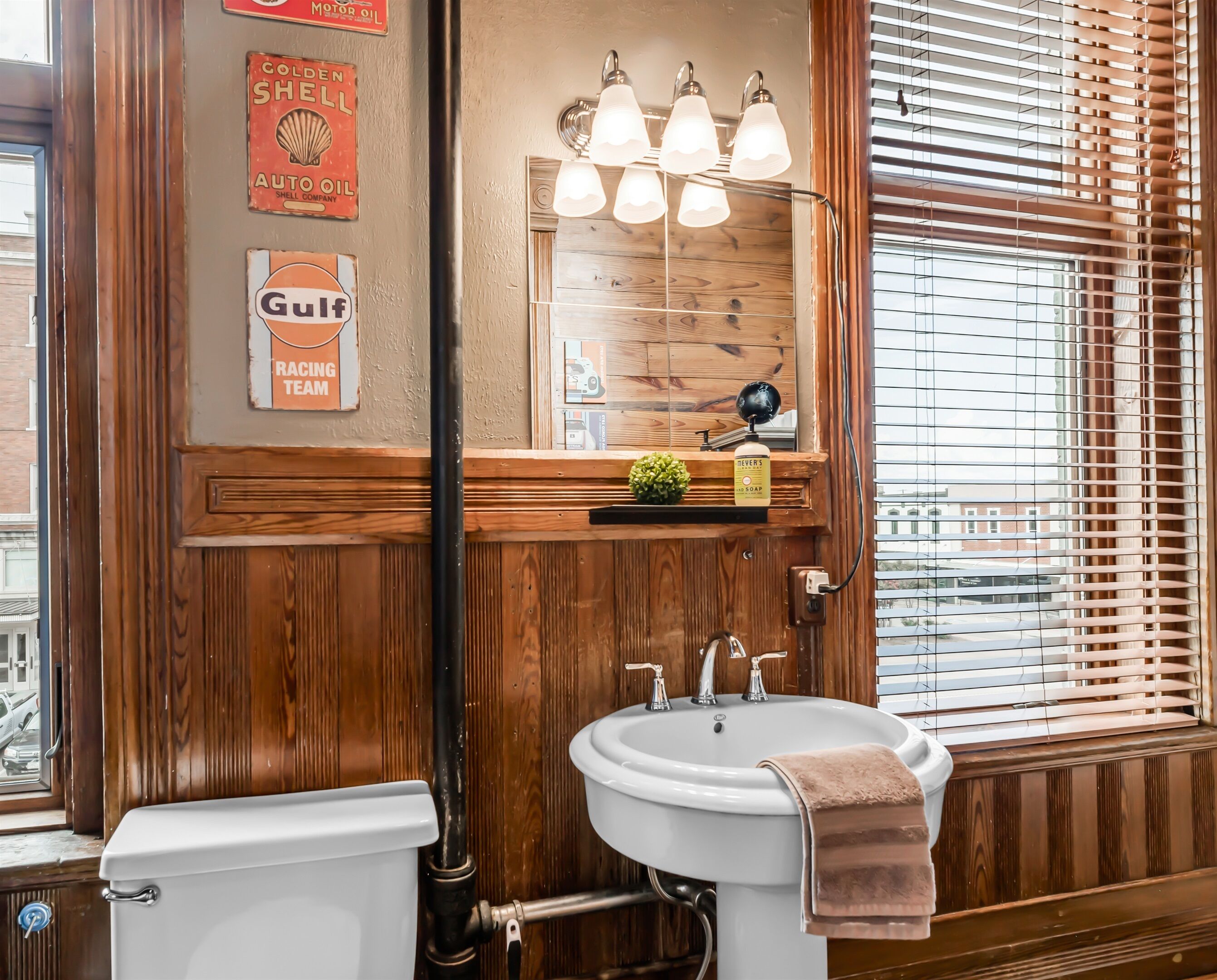 Une salle de bain avec un meuble-lavabo en bois, un lavabo blanc et des toilettes. On y trouve des affiches anciennes accrochées au mur, un miroir avec trois lampes et une fenêtre avec des stores.