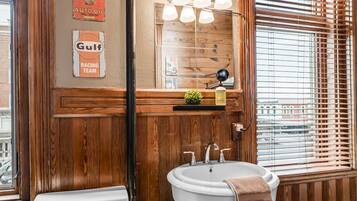 Une salle de bain avec un meuble-lavabo en bois, un lavabo blanc et des toilettes. On y trouve des affiches anciennes accrochées au mur, un miroir avec trois lampes et une fenêtre avec des stores.