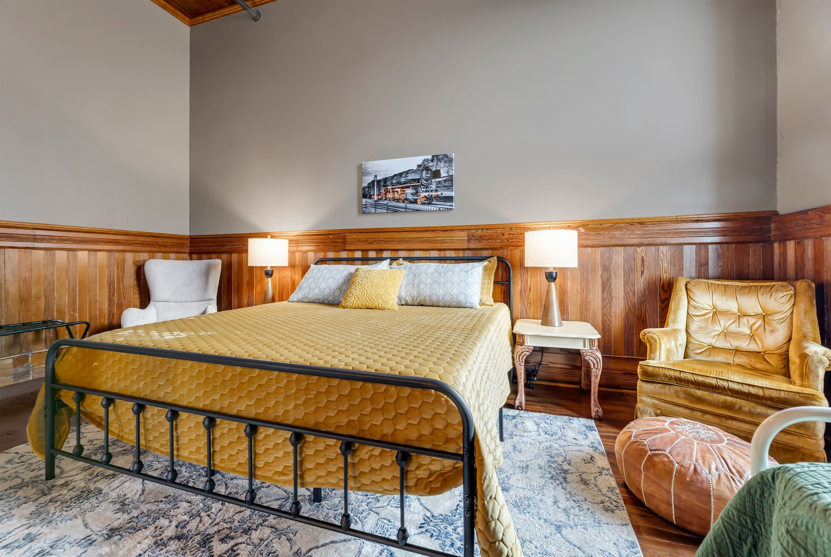 A bedroom with a bed, wooden headboard, two bedside lamps, a chair, a rug, and a painting on the wall.