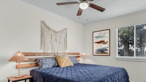 Une chambre avec une tête de lit en bois, une couvre-lit matelassée bleue, deux lampes de chevet, un tapis tissé, un ventilateur de plafond, une décoration murale en macramé et un tableau abstrait encadré.