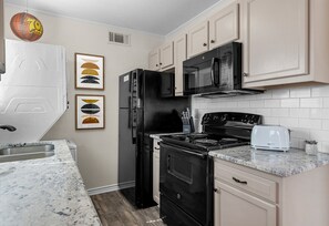 Una cocina con refrigerador negro, estufa y microondas, gabinetes blancos y encimera de mármol.