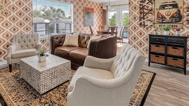 Un salon avec un mur orné d’un motif, un canapé en cuir marron, un fauteuil blanc, une table basse à motifs et un buffet agrémenté de paniers.