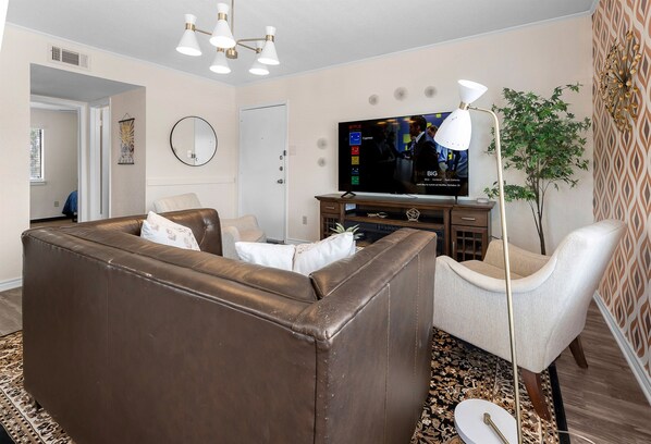 Una sala de estar con un sofá de cuero marrón, un sillón blanco, un televisor de pantalla plana, una lámpara de pie y una alfombra estampada.