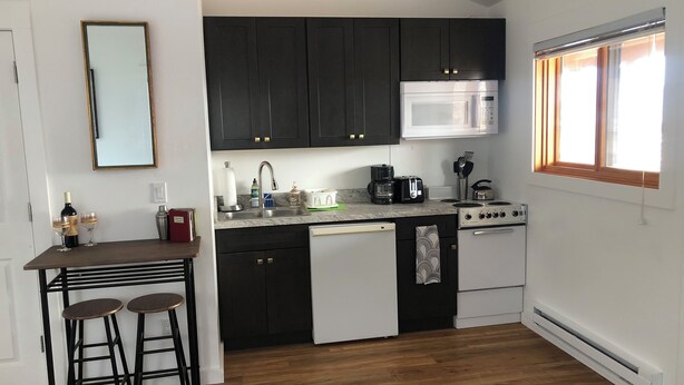 A compact kitchen with dark cabinets, a white microwave, a coffee maker, and a gas stove. There are two stools at a small table, and a window provides natural light.