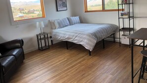 A bedroom with a bed, a desk, a chair, and two windows with a view of the countryside.
