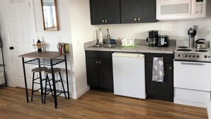 A compact kitchen with dark cabinets, a white microwave, a coffee maker, and a dishwasher. There is a small bar stool and a wooden table with a bottle and glasses.