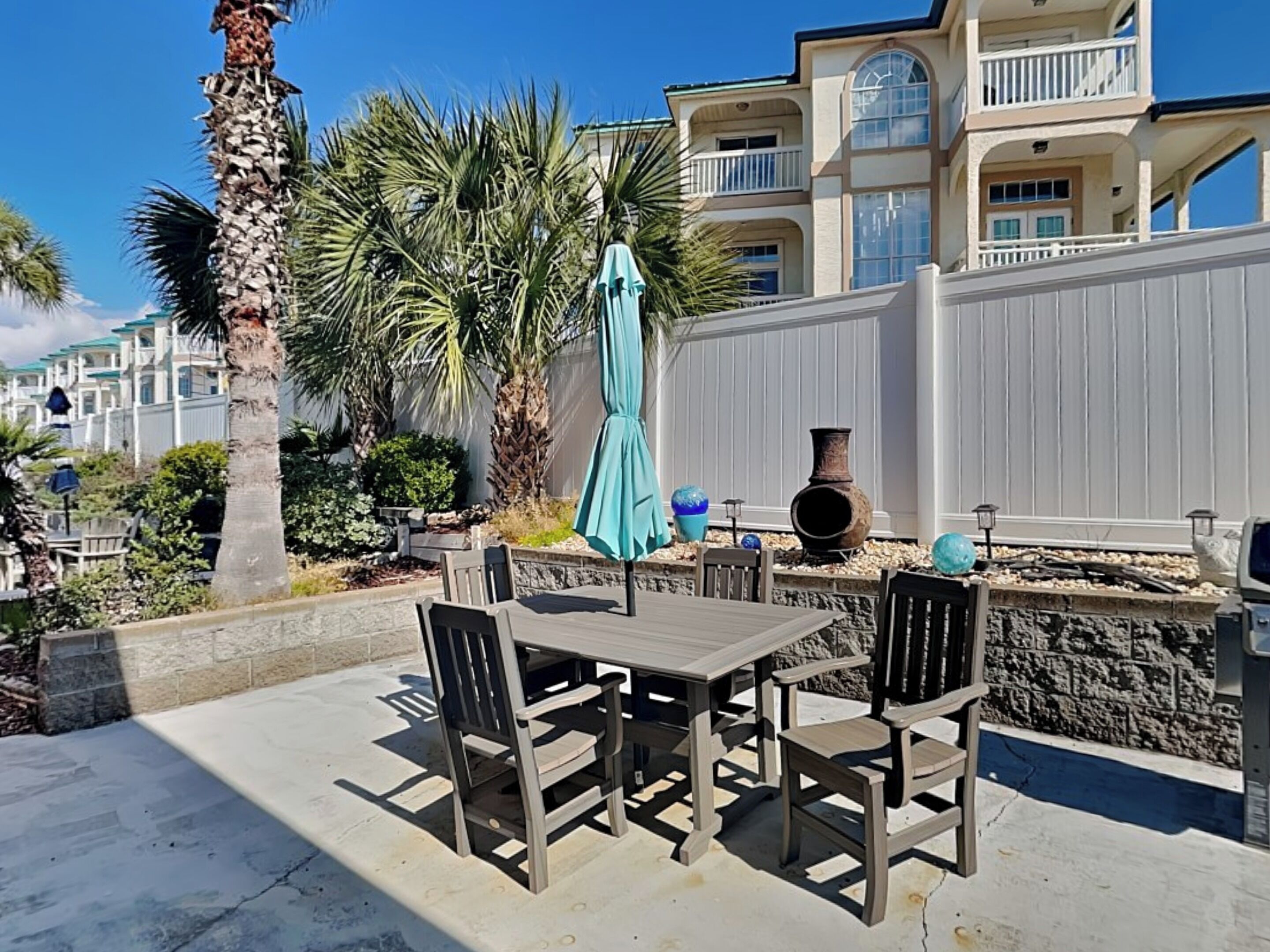 An outdoor patio with a table and chairs, surrounded by palm trees and a white fence.