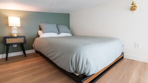 Un lit simple avec une couvre-lit gris clair, une table de chevet avec une lampe et un luminaire fixé au mur.