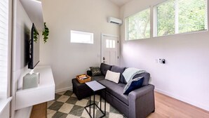 A compact living room with a sofa, a small table, and a TV mounted on the wall. There is a window with blinds, an air conditioning unit, and a door. - Extended Stay Retreat: Cozy yet Spacious Tiny Gem! (Austin)