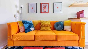 Een helder oranje bank met meerdere kussens, een rode poef, en ingelijste speelkaarten aan de muur.