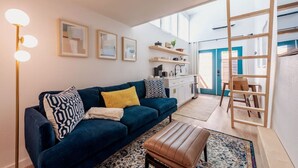A modern living room with a dark blue sofa, a wooden ladder, and a kitchen area visible through an open door. - Cozy & Bright Mid-Century Gem in Eastside Action! (Austin)