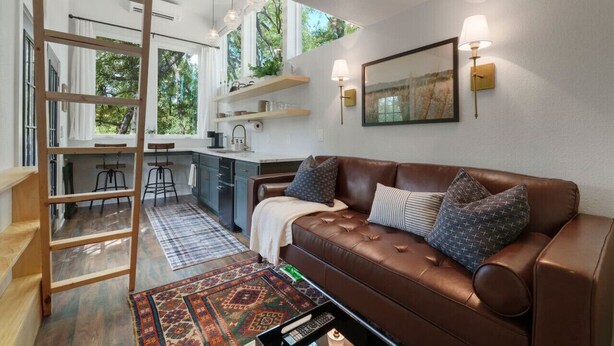 Una sala moderna con un sofá de cuero marrón, una escalera de madera, una zona de cocina y un amplio ventanal que proporciona luz natural.