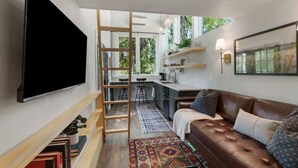 Una sala moderna con un sofá de cuero marrón, un televisor de pantalla plana montado en la pared, una escalera de madera y una zona de cocina con taburetes de bar.