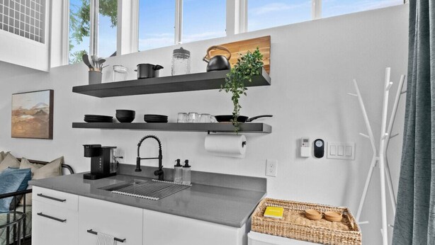 A modern kitchen with white cabinets, a black countertop, and a built-in oven. There are shelves above the sink holding kitchenware and a potted plant.