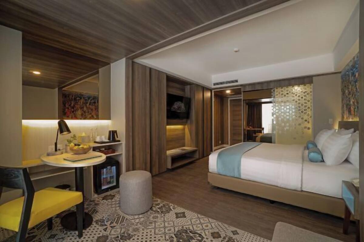 A modern hotel room with a bed, a desk with a lamp, a chair, and a small ottoman.