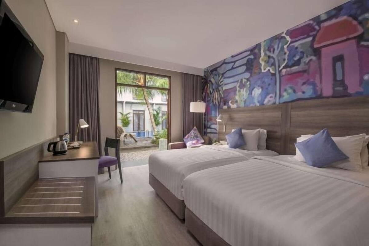 A hotel room with two beds, a desk, a TV, and a large window with a view of greenery.