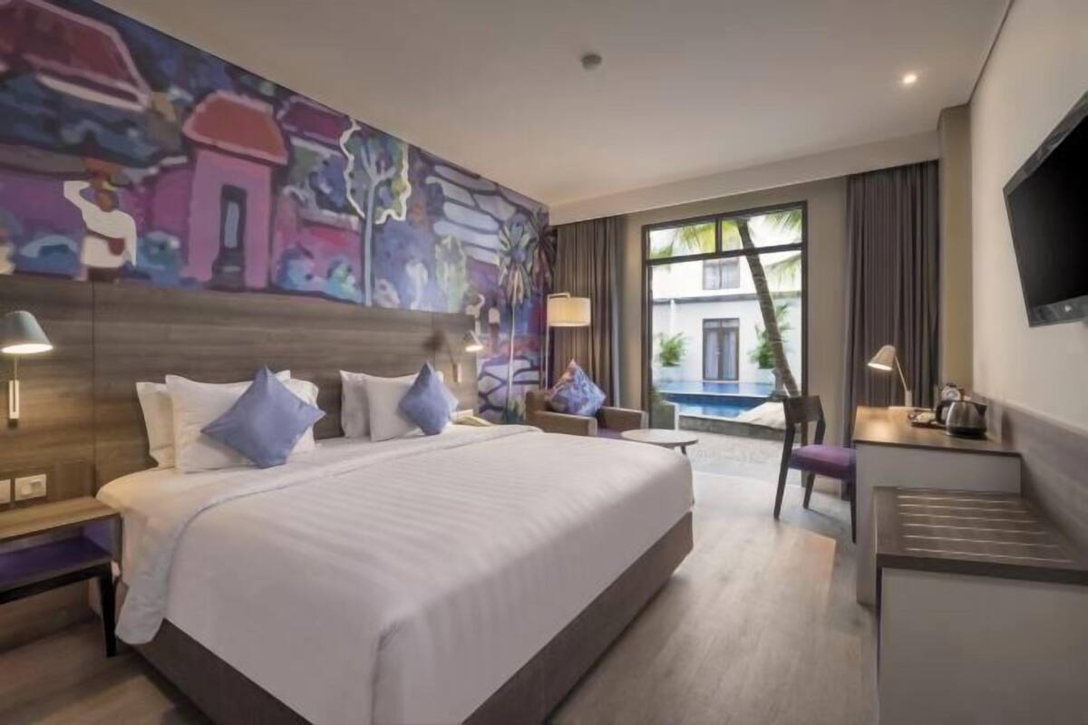A hotel room with a large bed, a desk, a chair, a TV, and a window with a view of a building and a pool.