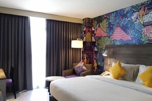 Quarto de hotel com cama, uma cadeira, uma escrivaninha e um mural colorido na parede.