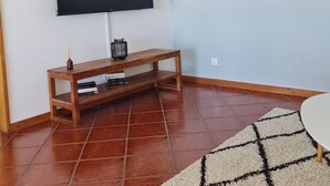 Una sala de estar con un televisor de pantalla plana montado en la pared, un mueble bajo de madera con libros y una vela, un tapete estampado y decoración en la pared.