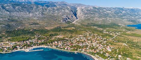 An aerial view of a coastal town with a mountainous backdrop, featuring a mix of residential buildings and open green spaces.