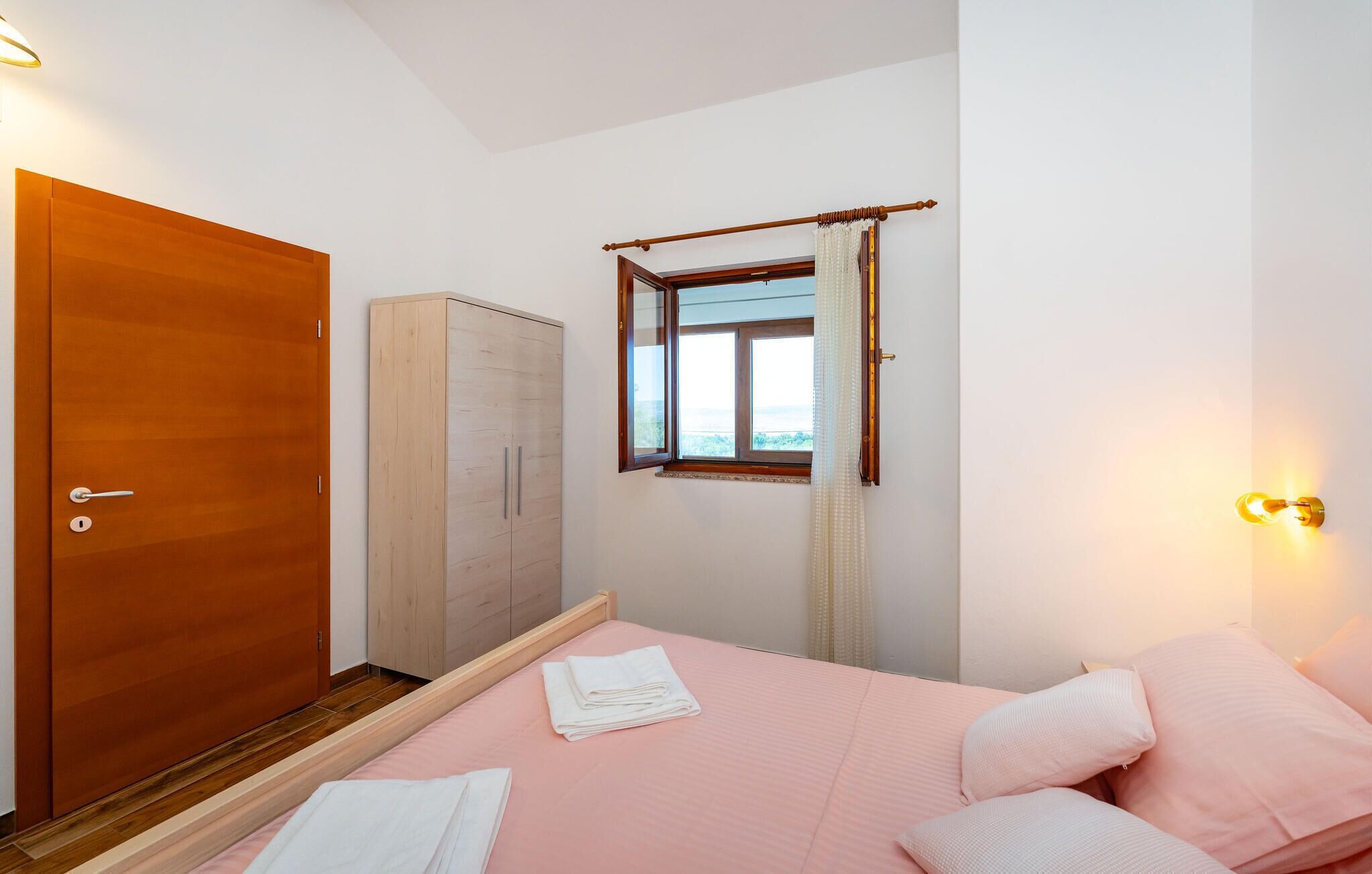 Une chambre avec une armoire en bois, une autre armoire avec des portes coulissantes, un lit avec une literie rose et une fenêtre donnant sur un beau paysage.