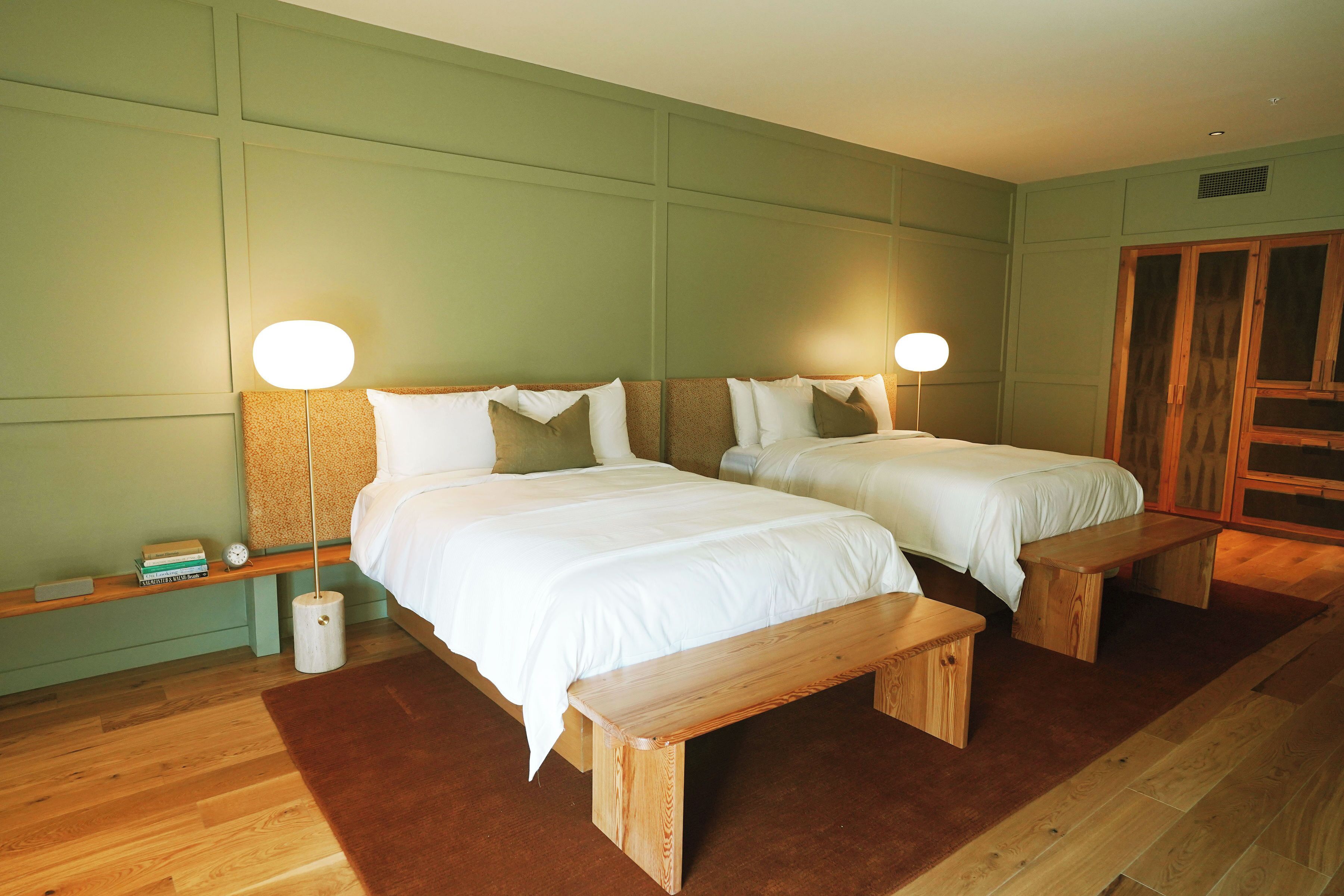 Habitación doble con dos camas, bancos de madera, una mesita de noche pequeña y una mesita auxiliar.