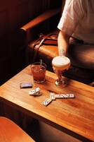 Een houten tafel met een drankje, dobbelstenen en een afstandsbediening.