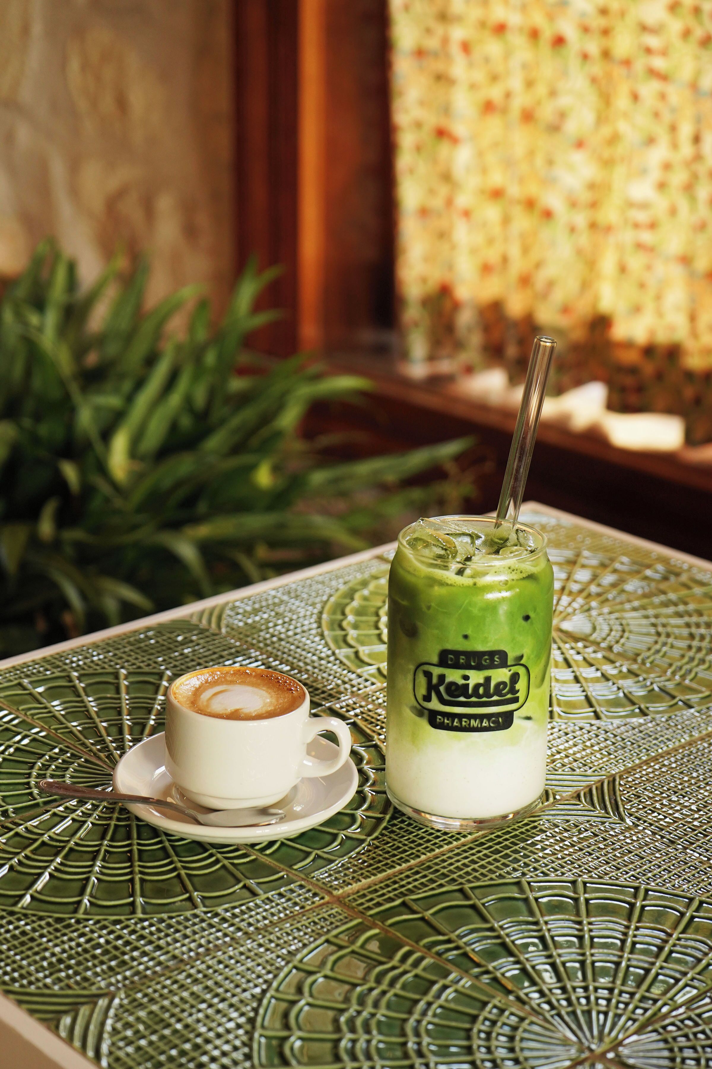 Una mesa con un mantel verde estampado, una taza blanca con platito, y una bebida verde con pajita.
