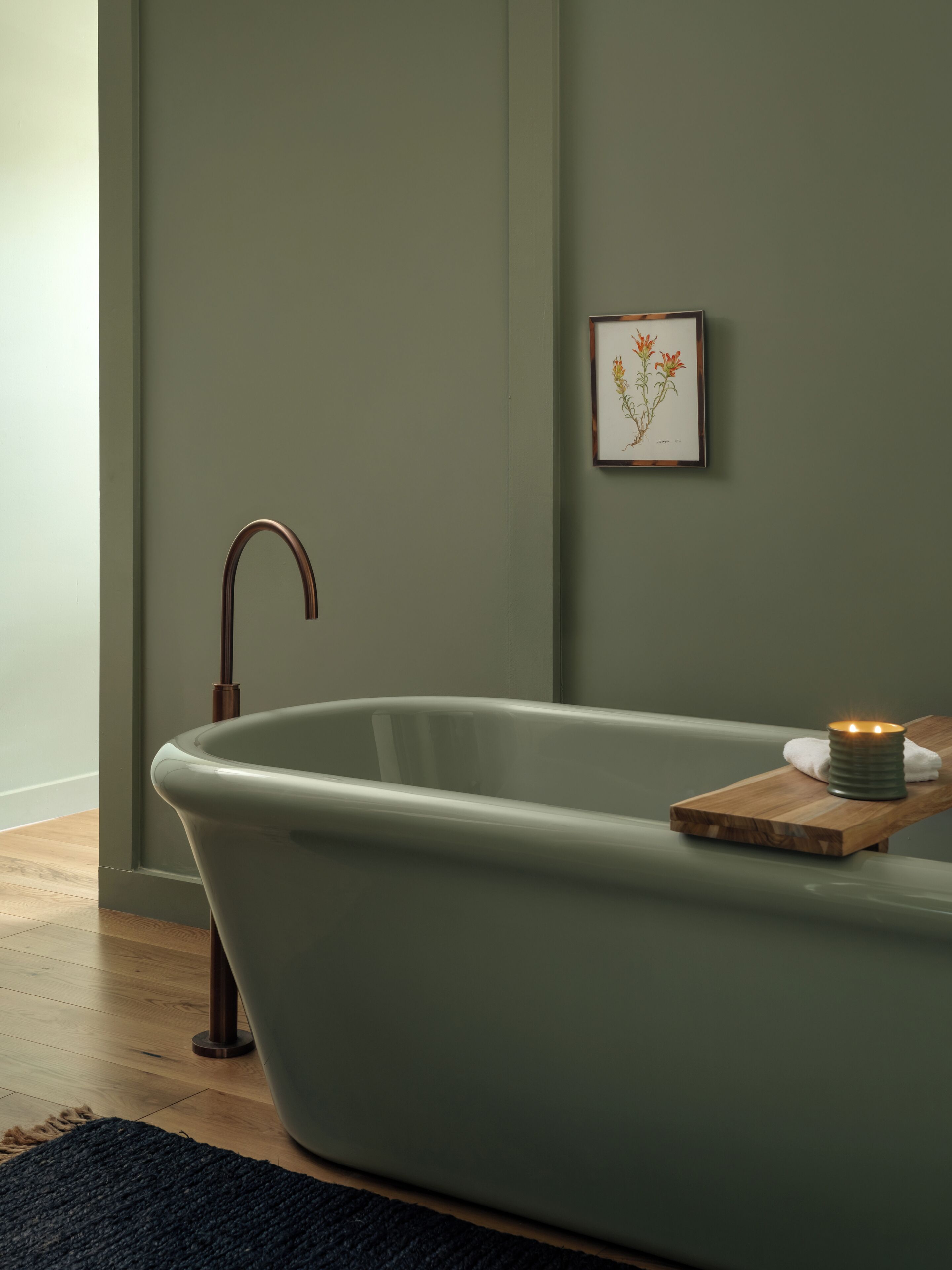 A modern bathroom with a freestanding bathtub, a framed botanical illustration on the wall, and a candle on a wooden tray.