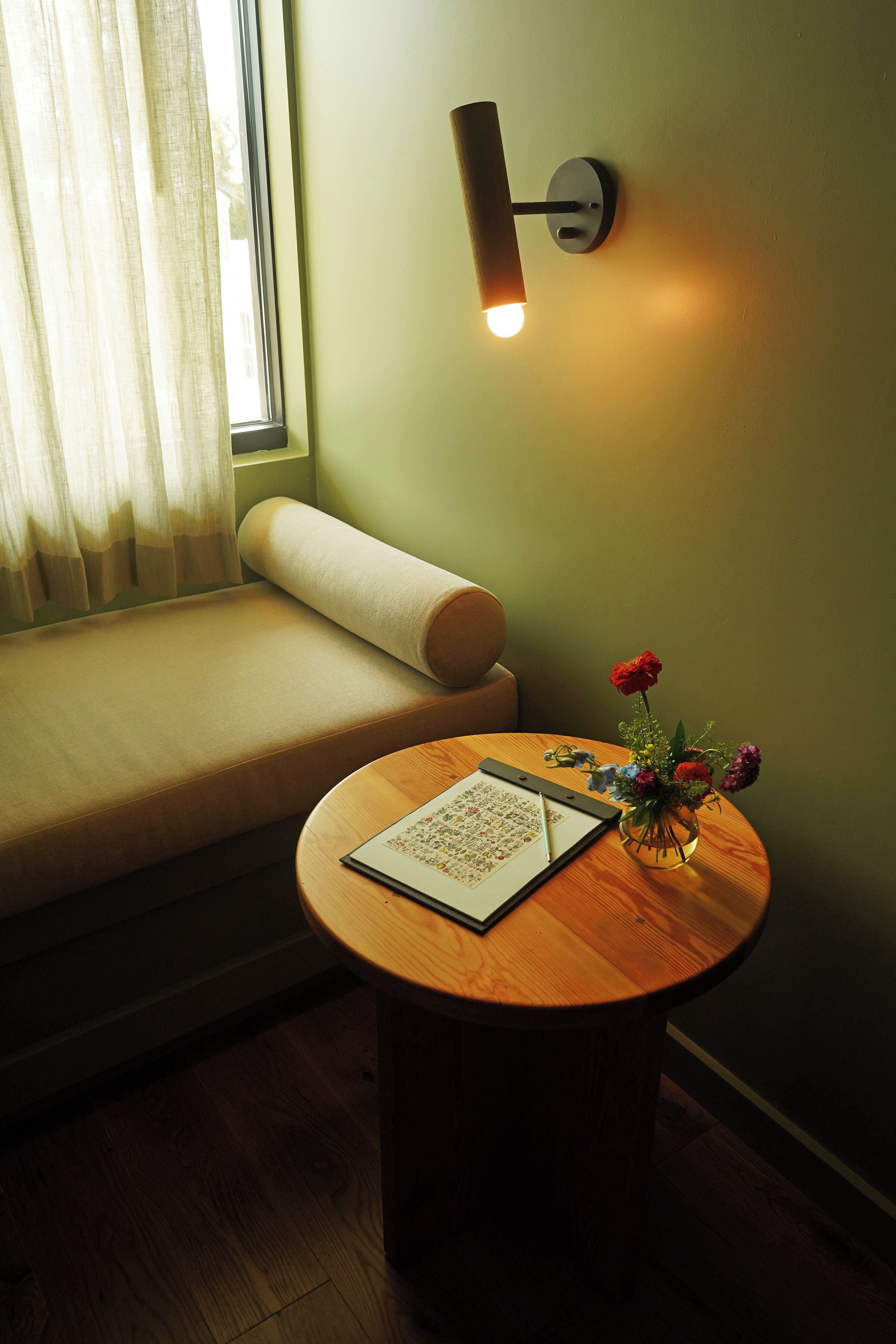 Una habitación acogedora con un asiento junto a la ventana, una mesa redonda de madera y un florero, y una lámpara fijada a la pared.