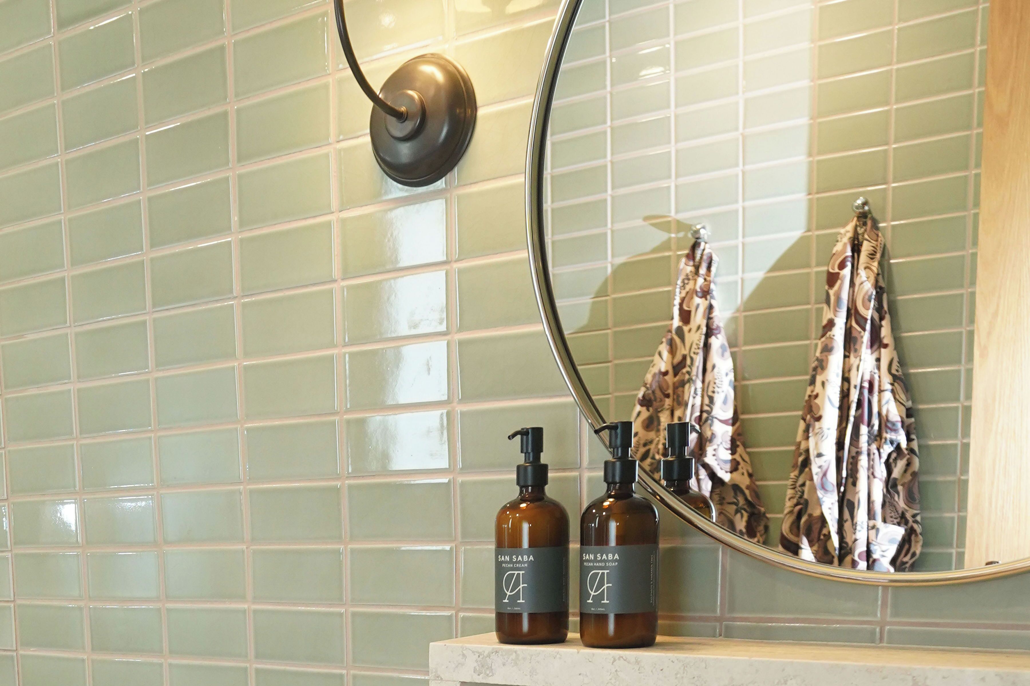 A bathroom with green tiled walls, a mirror, and two bottles of San Saba hand soap on a marble countertop.