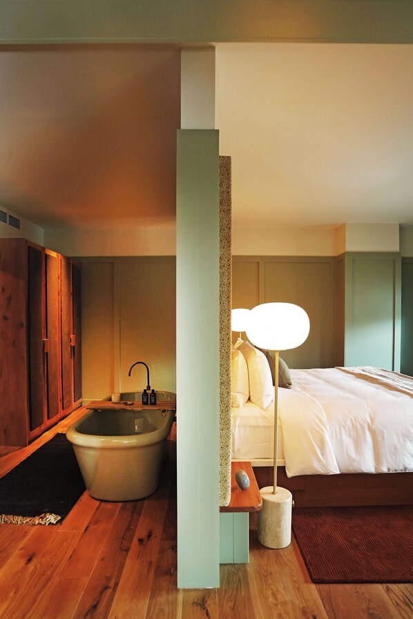 A modern hotel room with a wooden floor, a large bed, a bedside table with a lamp, and a bathtub. - Albert Hotel (Fredericksburg)