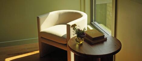 A room with a wooden floor, a white armchair, a small round table with a vase and books, a wall-mounted light, and a window with a view.