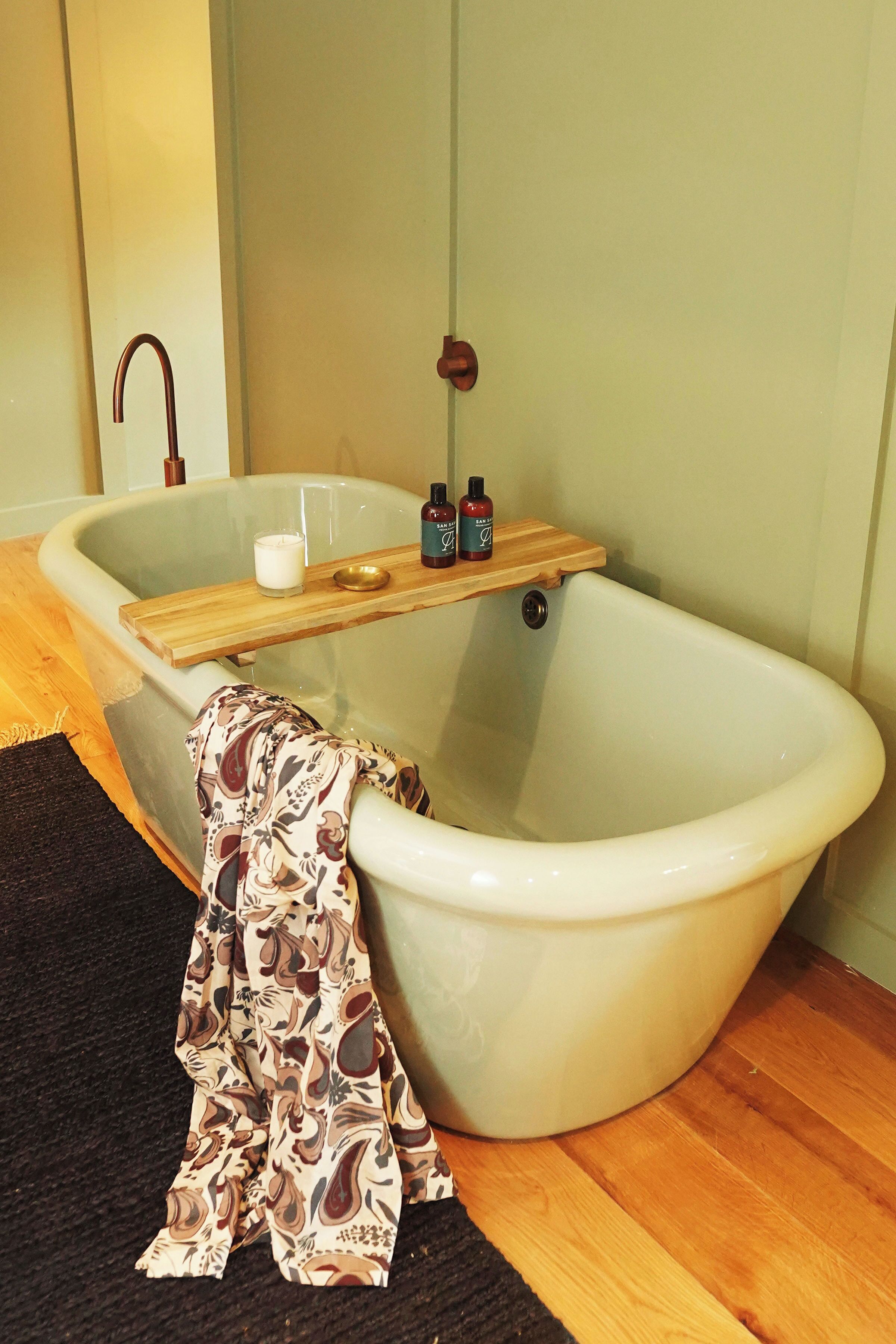 A bathroom with a freestanding bathtub, a wooden shelf with toiletries, and a patterned cloth on the floor.