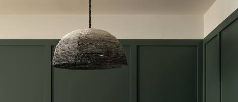 A dining area with a wooden table and chairs, a woven pendant light, and a dark green wall.