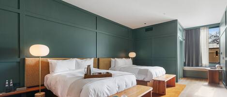 Quarto de hotel moderno com duas camas, abajures, um banco de madeira, uma mesa pequena e uma janela com cortinas.