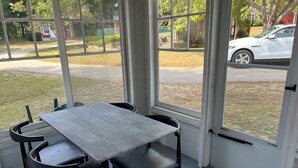 Una habitación iluminada por el sol con una mesa y sillas, ventanas amplias y vistas a la calle y áreas verdes.